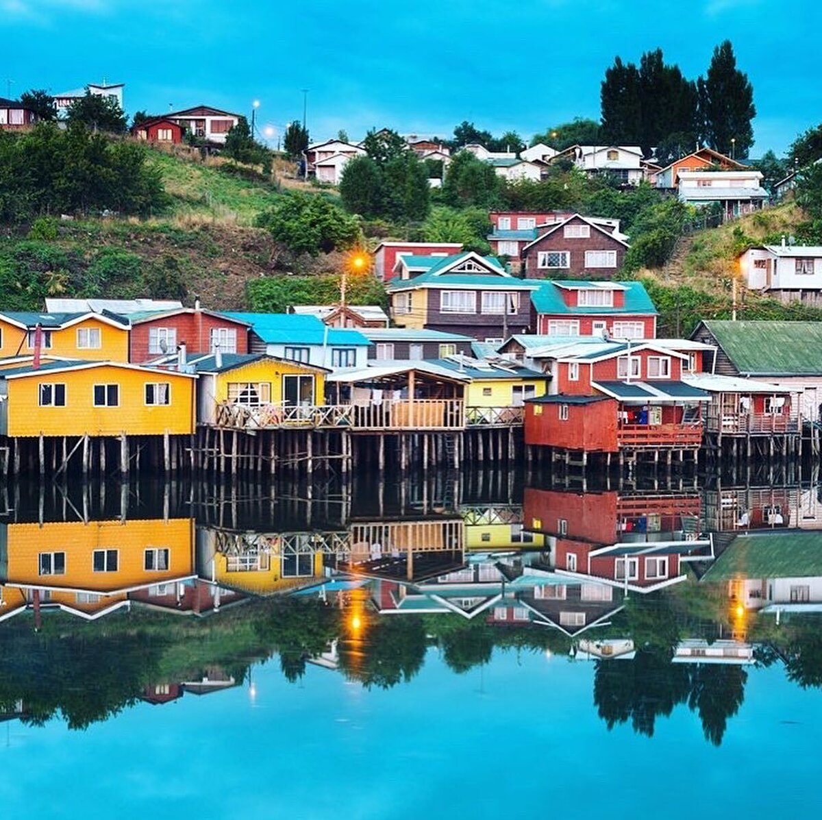 💐 Tek katlı müstakil ev görünümündeki kimi palafitoların önlerinde bulunan küçük balkon bahçelerdeki çiçekler göze çarpıyor hemen. Çiçeklerini sularken ettikleri derin sohbetle yılların komşuluğunu pekiştiren yaşlı kadınların renkli gülüşmeleri karışıyor sokağın ruhuna. Bu sohbet, evlerdeki kedilerin pencerelerden sokağa meraklı bakışlarıyla da karışınca sokağı adeta bir yağlı boya tabloya dönüştürüyor.⠀ Palafitolar arasındaki nadir bir boşluktan su kıyısına inildiğinde küçük sandallarında ağlarını temizleyen balıkçılarla karşılaşılıyor. Acelesiz, sakin bir hayat dönüyor palafitoların sütunları etrafında. Aklımıza ihtiyar bir balıkçının sözleri geliyor: “Chiloé’de bir söz vardır. Acele eden, zaman kaybeder. Yavaşlamak lazım…’’ 🌿⠀
