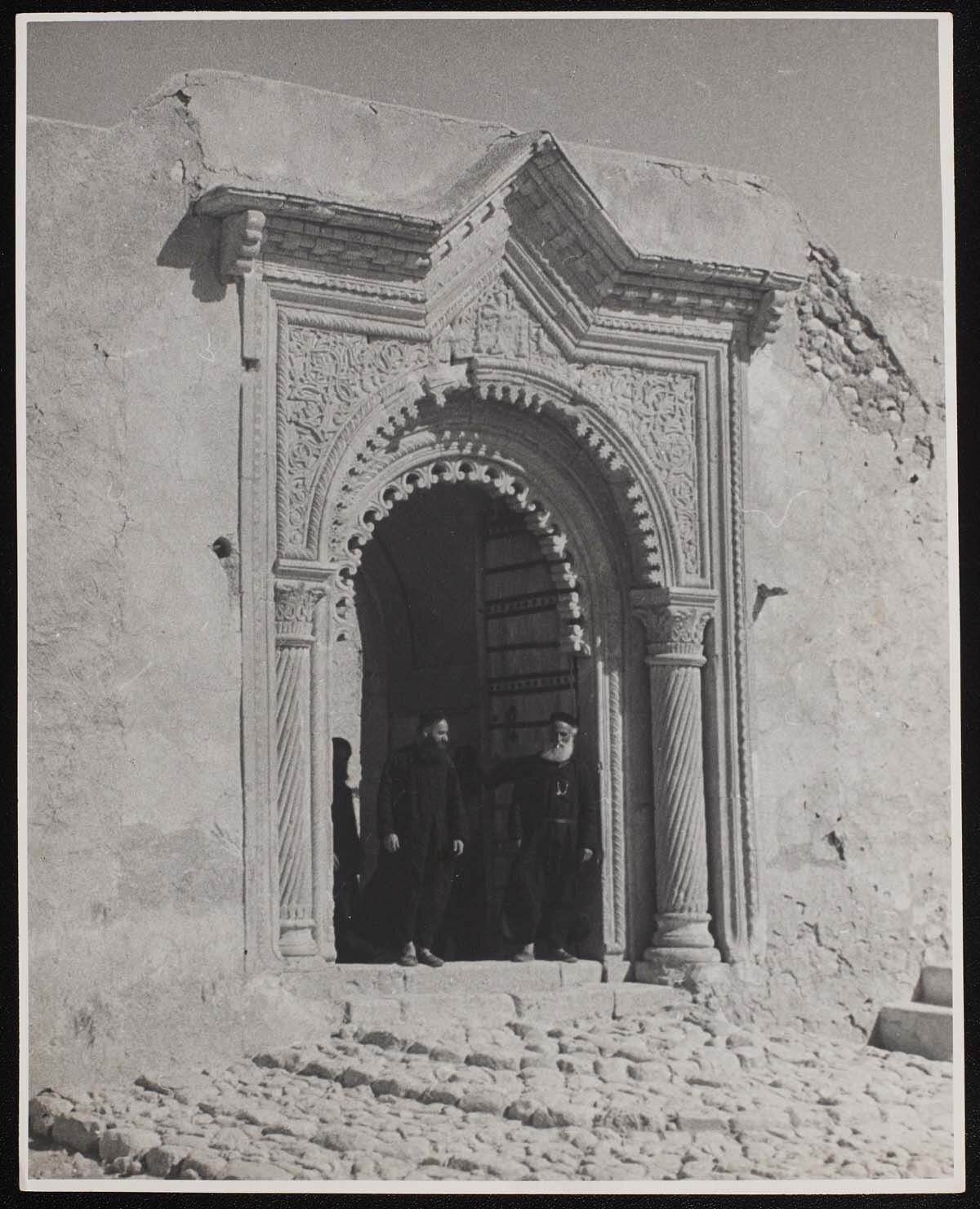 Alkuş Keldânî Manastırı'nın kapısı, Ağustos 1944. <br><br>Our Lady of the Seeds Manastırı, Alkuş'u çevreleyen dağlarda 640 yılında kurulan yakındaki Rabban Hormizd Manastırı'na modern ve daha merkezî bir alternatif olarak 1858'de inşa edilmişti. Bu fotoğraf, Kersting'in hem mimariye hem de bölgede yaptığı seyahatlerde karşılaştığı insanlara olan ilgisini gösteren birçok fotoğraftan biri.