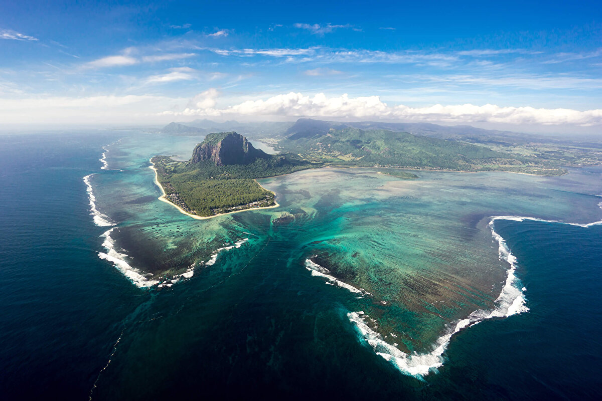 Sualtı Şelalesi - Mauritius<br><br>Sualtı Şelalesi, Mauritius’un güneybatısında, Le Morne Brabant Dağı’nın kıyısında yer almaktadır. Adanın muazzam berrak turkuaz suları, derin bir uçurum oluşturur. Bu uçurum, sanki bir sualtı şelalesi varmış gibi, adeta bir nehir gibi gözükür. Ancak, aslında burada akan bir su değil, kum ve tortu bulutlarıdır. Suyun yerçekimi etkisiyle aşağı doğru hareket etmesi, bu görkemli optik yanılsamayı yaratır. Bu etkileyici doğal oluşum, yerel halk arasında “Sualtı Şelalesi” olarak bilinir.