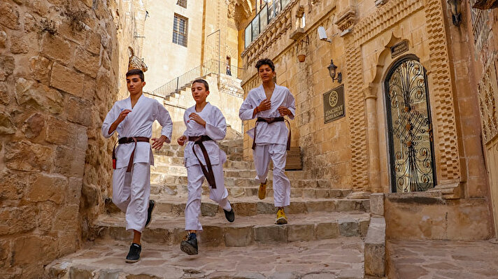 Koronavirüs tedbirleri kapsamında, 20 yaş altındakiler için getirilen 13.00- 16.00 saatleri dışındaki sokağa çıkma kısıtlamasında Mardin'deki genç karateciler Enes Ömer Yıldızoğlu (14), Hüseyin Çaça (13) ve Abdullah Can (13), gelecek aylarda yapılacak milli takım seçmelerine hazırlanıyor. Genç karateciler, bu süreçte çalışmalarını hem evlerinde hem de kısıtlamadan muaf oldukları saatlerde, antrenörleri Murat Kaya eşliğinde kentin tarihi ve dar sokaklarında antrenman yaparak sürdürüyor. Uzun süre evde kalmaları nedeniyle çok basamaklı merdivenlerde koşu yapan karateciler, hız, kuvvet ve dayanıklılığa yönelik antrenmanlarla milli takıma girmek için ter döküyor. 