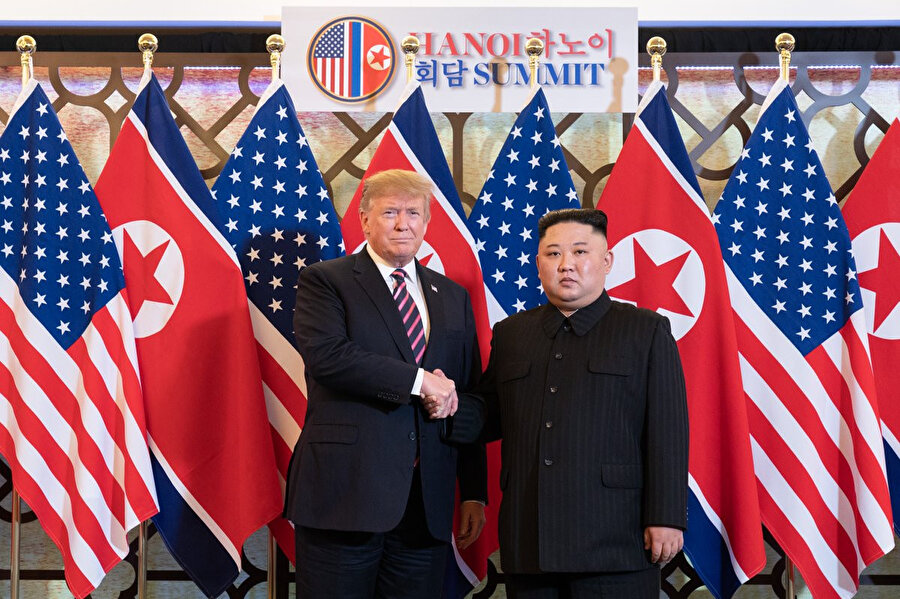Donald Trump ve Kim Jong ülke bayraklarının önünde el sıkışırken görünüyor.
