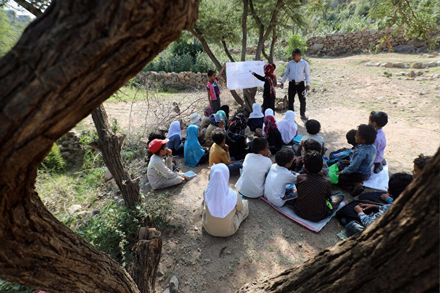 16 Eylül 2019’da Yemen’in Taiz vilayetine bağlı bir köyde çekilen bu fotoğrafta henüz inşaatı tamamlanmamış okullarının yakınında bir ağacın gölgesinde ders işleyen çocuklar. 