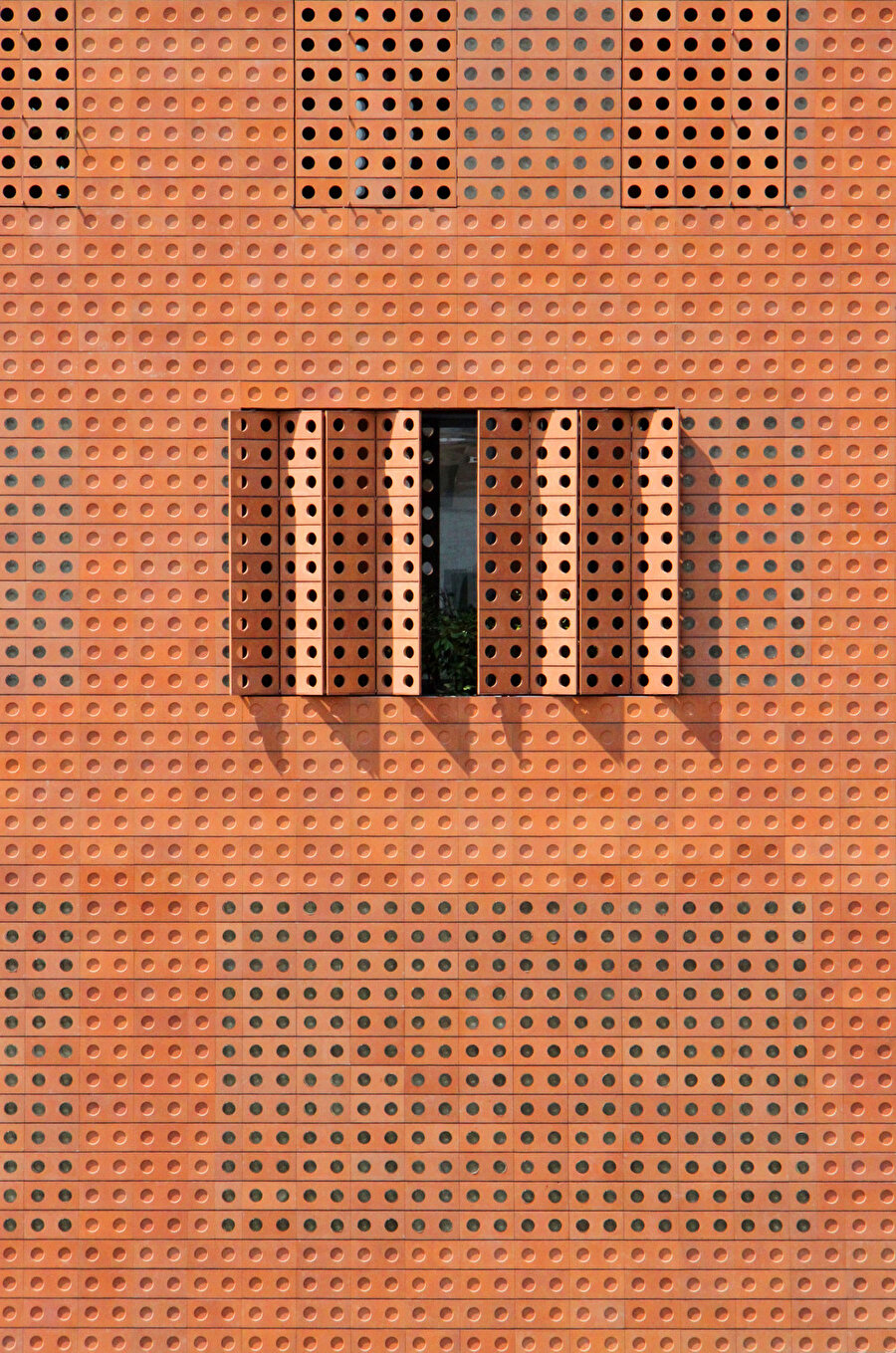  Tuğla ile aynı pas rengi tonundaki ve uyumlu dairesel oyuklara sahip akordeon panjurlar, cephe için birleşik bir görünüm oluşturmak için binanın pencerelerini kaplıyor. 