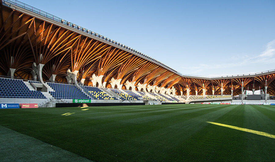 Bu fotoğraf hakkında bir şeyler yazAhşap çatı strüktür, stadyumun iç mekan kimliğini tanımlıyor.