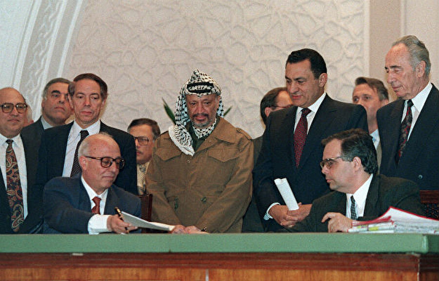 Filistin Kurtuluş Örgütü'nden Filistinli arabulucu Ahmed Kurey (masada solda) ve Oslo Barış Anlaşmalarının baş mimarları İsrailli diplomat Uri Savir (masada sağda), 09 Şubat 1994'te, Kahire'de, Gazze ve Eriha'daki Filistin özyönetimi konusunda varılan anlaşmaları imzalarken FKÖ başkanı Yaser Arafat (ayakta ortada), Mısır Devlet Başkanı Hüsnü Mübarek (sağda 2) ve İsrail Dışişleri Bakanı Şimon Peres (sağda) oradaydı.