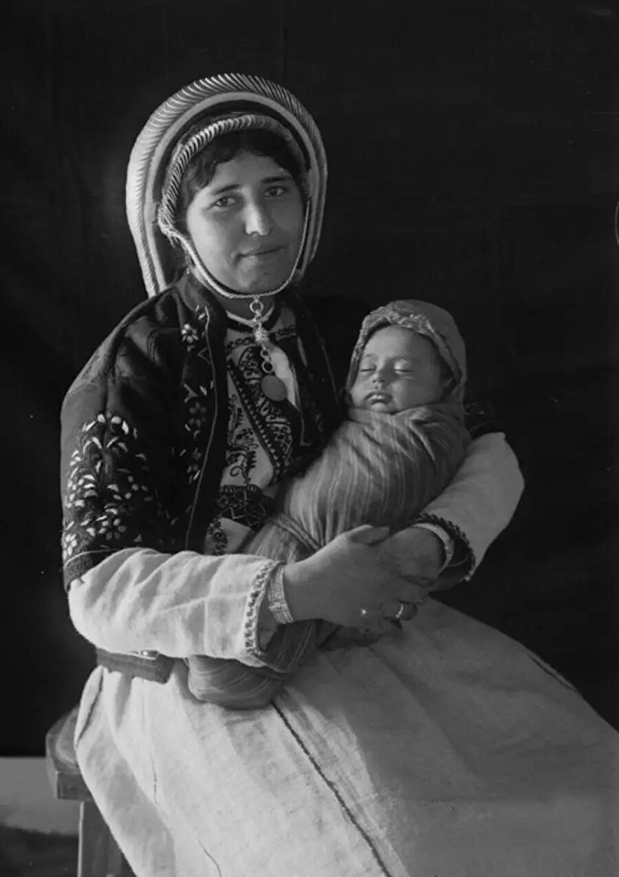 Ramallah’tan geleneksel işlemeli elbise giyen Filistinli bir kadın, kucağındaki bebeğiyle poz veriyor, 1920'ler-30'lar.