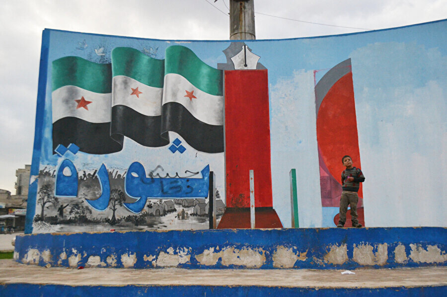 Şehir merkezindeki anıtın üzerinde özgür Suriye’yi temsil eden bir bayrak, kalem ve kılıç bulunuyor. Bayrağın hemen altında sevra (devrim) yazıyor.