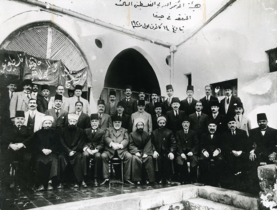 Filistin'in ana şehirlerini ve bölgelerini temsil eden Üçüncü Filistin Ulusal Kongresi delegeleri. Pankartta sağdan sola Arapça şöyle yazıyor: "Filistin Hz. İsa'nın beşiğidir"; "Mescid-i Aksâ'yı koruyun"; "Filistin Araptır." Son sıranın sağdan üçüncüsü, müstakbel Filistin lideri Hacı Emîn el-Hüseynî, Kudüs müftüsü olmadan birkaç ay önce. Hayfa, 1920.