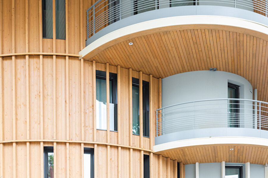 Pencere açıklıklarının ritmi ve ahşap kaplama detayları arasındaki hassas denge cephenin derinlik etkisini artırırken, beyaz bordürler yatay hatları vurguluyor.