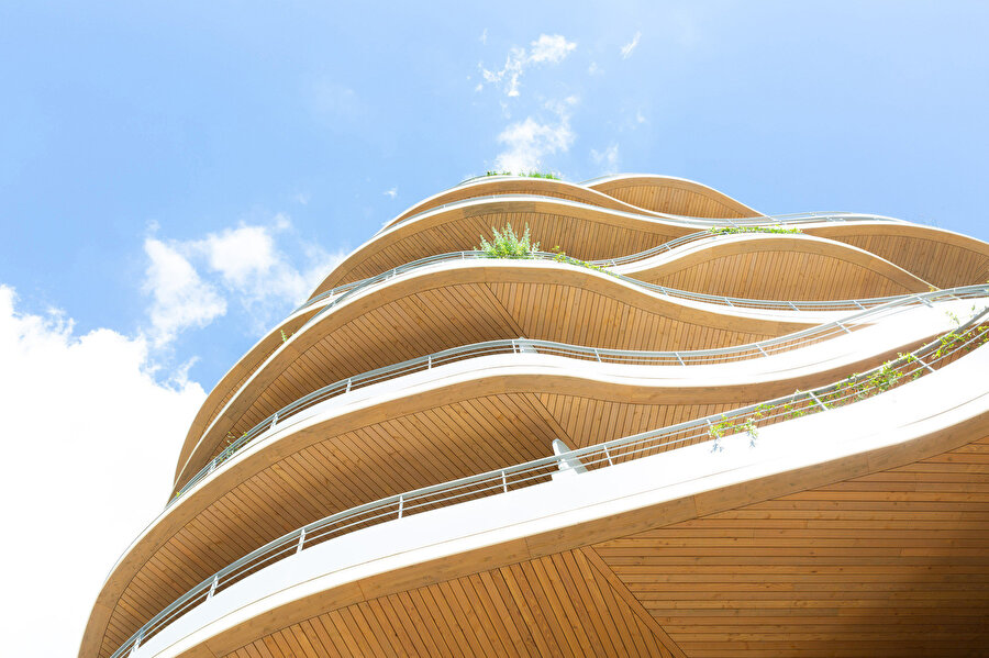 Ahşap kaplı tavan yüzeyleri ve beyaz balkon parapetleri arasındaki kontrast yapının dalgalı silüetini tanımlarken üst kotlarda konumlandırılan bitkiler gökyüzüyle yumuşak bir geçiş oluşturuyor.