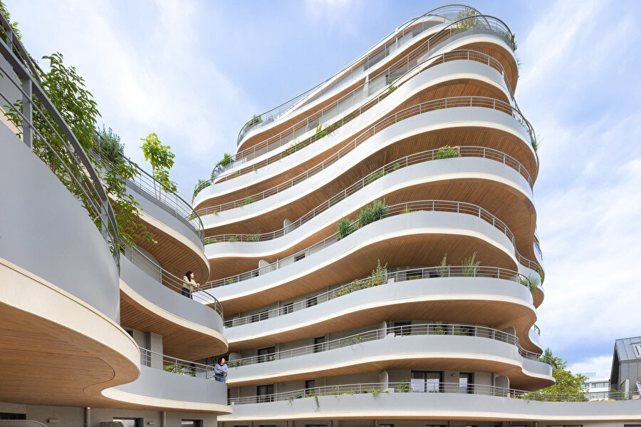 Her katta tekrar eden eğrisel balkonlar ve yeşil bitki öğeleri dikey bahçe etkisi oluştururken ahşap tavan kaplamaları yapıya sıcak bir karakter kazandırıyor.