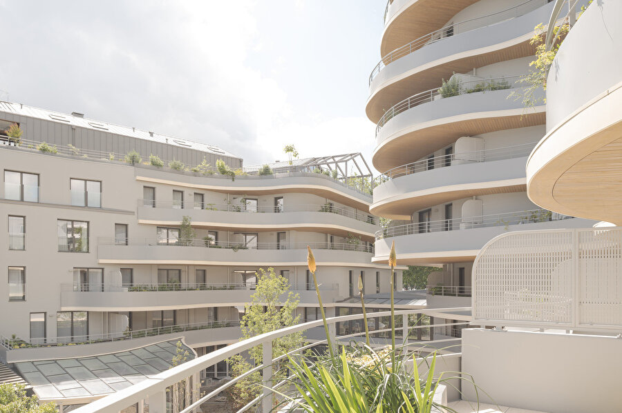 Terasların kademeli yerleşimi ve yeşil bitki öğeleri yapının farklı kotlarında doğal bir geçiş sağlarken eğrisel balkonlar binanın organik formunu vurguluyor.