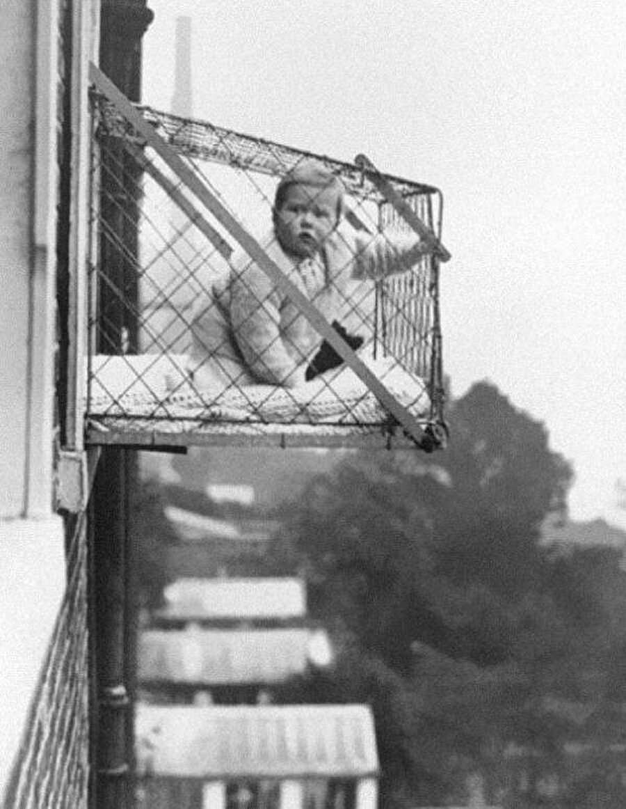 
                                    Fotoğraf 1922 yılına ait. 1920'li ve 30'lu yıllarda Londra'da çok popüler bir uygulamaymış, bebek kafesleri. Yüksek binalarda yaşayan aileler, bebeklerinin güneş ışığından daha çok yararlanabilmesi için pencerelerine bu aparatı yerleştiriyormuş.
                                