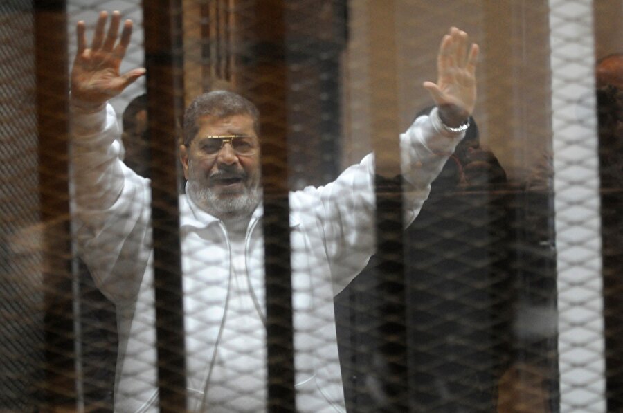 Darbeyle görevinden uzaklaştırılan Muhammed Mursi, 8 Ocak 2015 tarihinde Kahire'deki polis akademisinde görülen duruşma sırasında parmaklıkların arkasından mahkeme salonunda bulunanları selamlıyor.