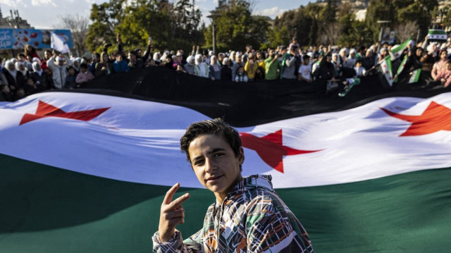 Un garçon fait un geste en posant devant des personnes agitant un drapeau syrien géant datant de l'époque de l'indépendance lors d'une célébration de l'éviction du président syrien Bachar al-Assad sur la place des Omeyyades dans le centre de Damas, le 13 décembre 2024.