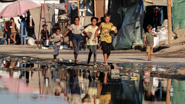 Des enfants palestiniens portant des conteneurs vides marchant près d'eaux usées stagnantes, alors qu'ils se rendent à un point de distribution de nourriture à Deir el-Balah, dans le centre de la bande de Gaza, le 19 juillet 2024. 