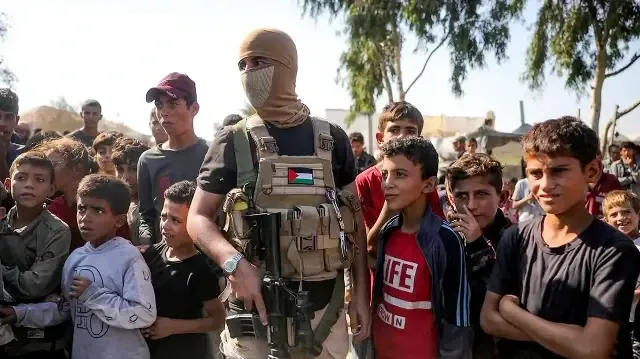 Un combattant palestinien des Brigades Ezzedine al-Qassam, la branche armée du mouvement palestinien Hamas, monte la garde à côté d'enfants avant l'arrivée des véhicules du Comité international de la Croix-Rouge (CICR) dans le sud de Deir el-Balah, dans le centre de la bande de Gaza, avant la remise du deuxième lot d'otages qui devraient être libérés, le 13 octobre 2025.