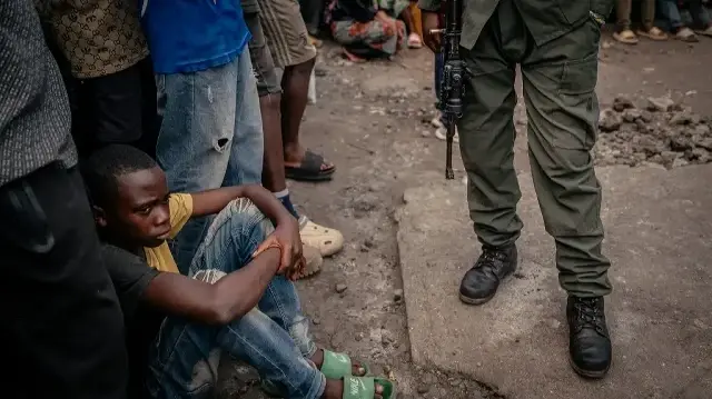 Un jeune garçon est assis par terre à côté d'un soldat de l'AFC/M23 tenant son arme, chargé d'assurer la sécurité de la réunion, à Goma, le 24 juillet 2025. Ce rassemblement s'inscrit dans le cadre d'une stratégie de sensibilisation visant à instaurer la confiance auprès de la population locale. 
