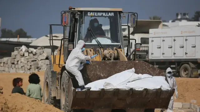 Une excavatrice transportant les corps de Palestiniens passe devant des enfants assis lors d'un enterrement collectif dans un cimetière de Khan Yunis, dans le sud de la bande de Gaza, le 10 novembre 2025.