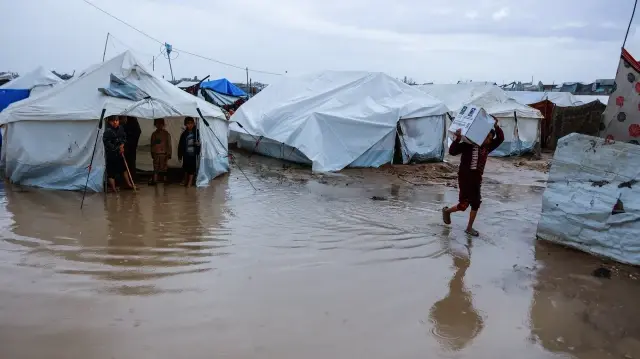 Un garçon transporte une boîte à travers une flaque d'eau dans un camp de fortune abritant des Palestiniens déplacés après de fortes pluies dans le quartier de Zeitoun, à Gaza, le 11 décembre 2025.