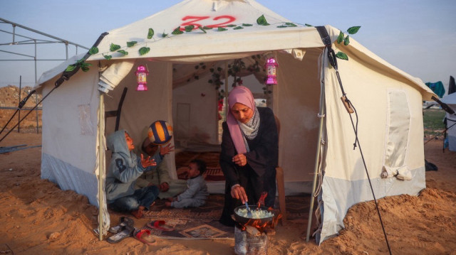 Une femme palestinienne prépare un repas tandis que des enfants sont assis à ses côtés, dans leur tente de déplacement au camp de réfugiés de Bureij, dans le centre de la bande de Gaza, le 5 mars 2025.