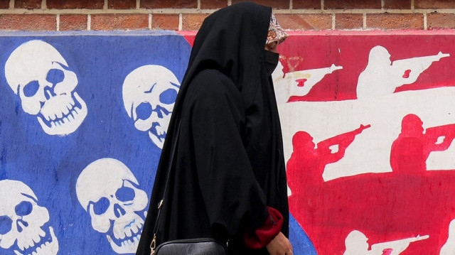 Une femme passe devant une fresque représentant le drapeau américain, dont les étoiles ont été remplacées par des crânes et les bandes blanches et rouges par des silhouettes de soldats tirant des fusils d'assaut, peinte sur les murs extérieurs de l'ex-ambassade des États-Unis à Téhéran, surnommée colloquialement le "Spy Den", le 8 avril 2025.