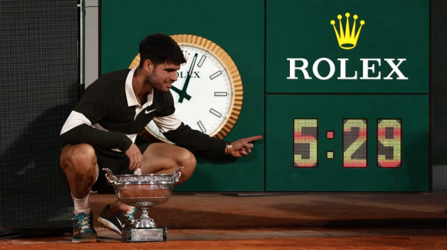 L’Espagnol Carlos Alcaraz pose avec le trophée à côté de l’horloge indiquant la durée du match, la plus longue jamais enregistrée pour une finale à Roland-Garros, après sa victoire contre l’Italien Jannik Sinner à l’issue de la finale messieurs du tournoi de Roland-Garros, sur le court Philippe-Chatrier, à Paris, le 8 juin 2025.