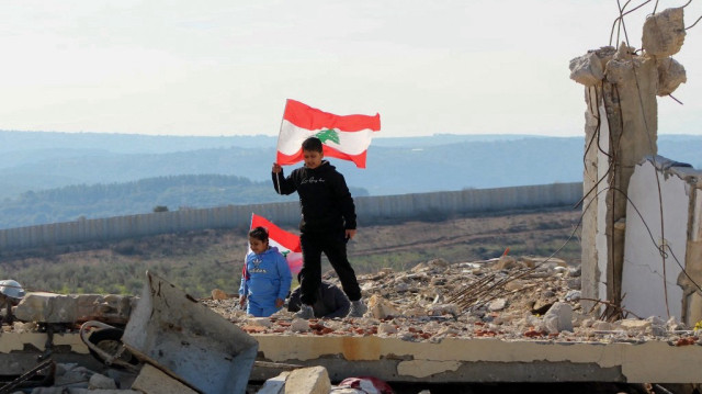 Des enfants libanais brandissent leur drapeau national alors qu'ils marchent au-dessus des décombres d'un bâtiment détruit, tandis que des familles retournent avec l'armée libanaise dans le village méridional de Marwahin, près d'une barrière frontalière avec Israël (à l'arrière-plan), le 28 janvier 2025.
