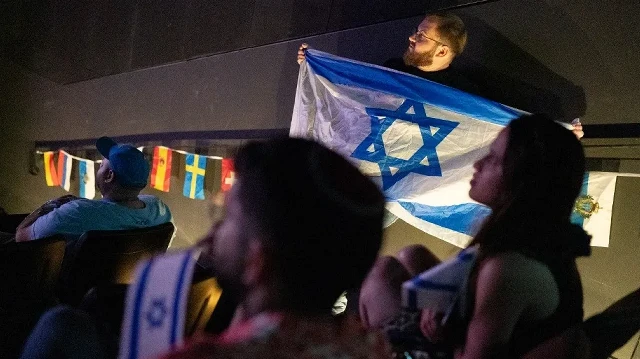 Un homme tient un drapeau israélien tandis que des personnes regardent la finale du Concours Eurovision de la chanson 2025 diffusée dans un centre communautaire à Tel Aviv, en Israël, tôt le matin du 18 mai 2025. 