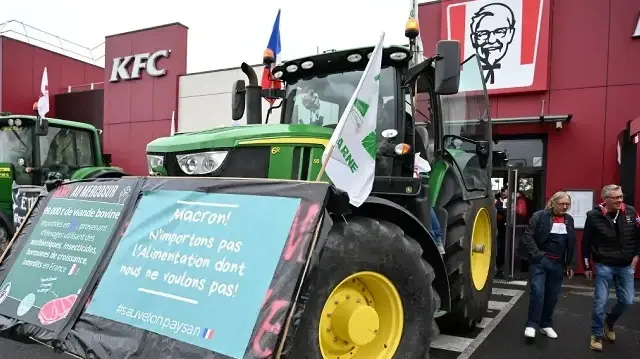 Des membres du syndicat agricole français FDSEA (Fédération départementale des exploitants agricoles) et des Jeunes Agriculteurs (JA) garent leurs tracteurs avec une pancarte sur laquelle on peut lire "Macron : N'importez pas les aliments dont nous ne voulons pas" devant un restaurant fast-food KFC lors d'une manifestation contre la concurrence déloyale des produits agricoles importés et l'accord de libre-échange avec le Mercosur, à Saint-Memmie, dans la banlieue de Châlons-en-Champagne, dans l'est de la France, le 26 septembre 2025. 