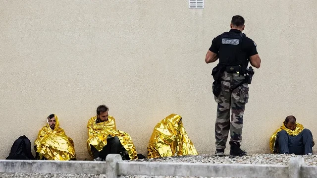 Un gendarme français se tient parmi des migrants abandonnés par un passeur, assis sous des couvertures de survie après avoir tenté de traverser la Manche au large de la plage d'Hardelot, à Neufchâtel-Hardelot, dans le nord de la France, le 13 août 2025. 