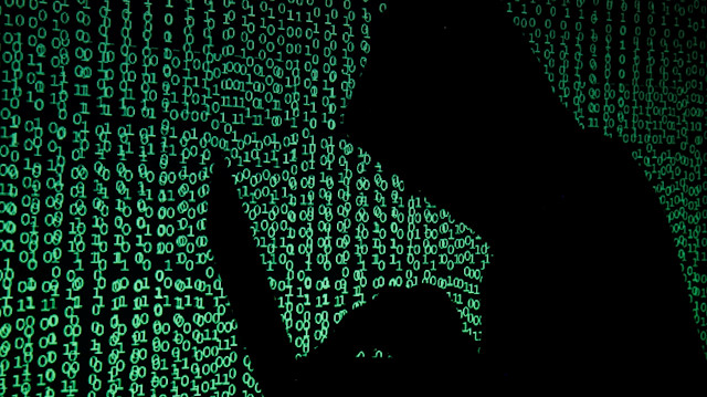 A hooded man holds a laptop computer as cyber code is projected