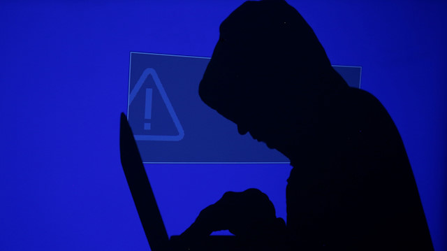 A hooded man holds a laptop computer as blue screen with an exclamation mark is projected 